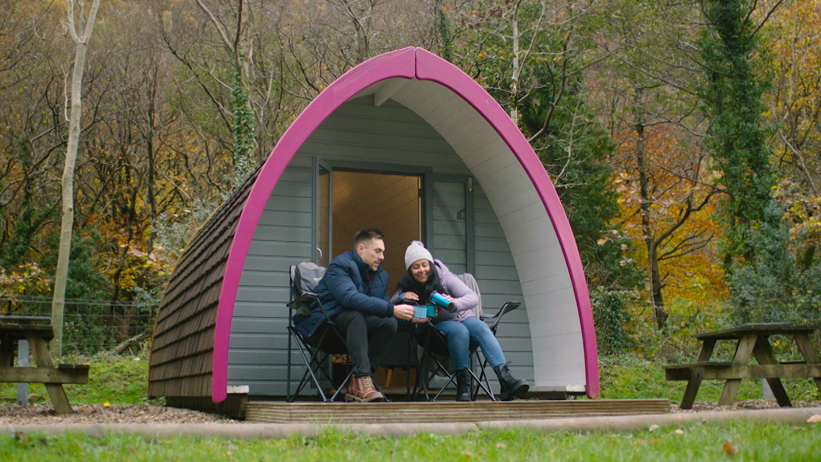 Woman pouring a drink into a mug held by a man both sitting down and are outside